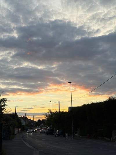 The image shows a street scene in Orsay, France, at dusk. The sky is filled with a dramatic cloudscape featuring hues of gray, white, and orange, suggesting a sunset. A street runs through the scene, lined with streetlights and power lines. On the right side of the road, there is dense foliage and parked cars. Further down the street, car headlights are visible, indicating traffic. Houses are visible in the distance. The image has a subdued, warm lighting, likely due to the setting sun.