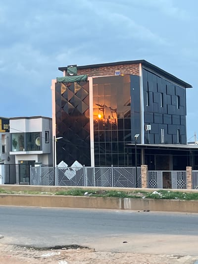 The image depicts a modern, multi-story building with a reflective, black glass facade, situated in Offatedo, Nigeria. The building's design features geometric patterns on its upper section. A green tarp is draped over a portion of the rooftop, obscuring some details. The sun is setting or rising, casting a warm glow and reflecting brightly on the glass, creating an impression of a large, orange orb. Adjacent to this building is a smaller, lighter-colored structure with large windows. In front of both buildings, a grey metal fence with intricate geometric designs encloses the property. A paved road runs along the foreground, with a concrete barrier and a strip of green grass separating it from the fenced area. The sky is overcast with light grey clouds. A few pieces of debris are scattered on the roadside and the grass verge.