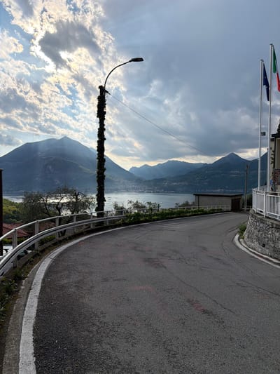 The image depicts a view in Gittana, Italy. The scene is rotated such that the road curves upwards on the right. The foreground features a road with a white line marking its edge and a metal guardrail. A light pole, heavily overgrown with vegetation, stands near the guardrail. Beyond the road and guardrail is a scenic view of a lake surrounded by mountains, partly obscured by clouds and sunlight. Flags (possibly Italian and European) are visible atop poles in the upper left. The sky is filled with clouds, with the sun breaking through in some areas.
