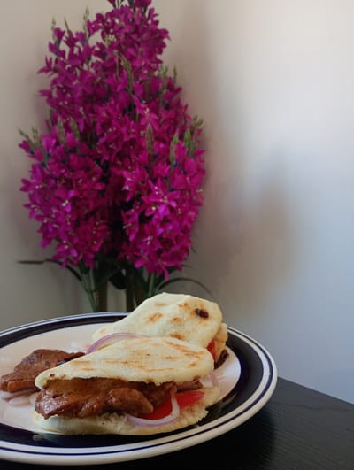 A close-up shot features a plate of food, likely a meal, arranged on a dark surface in an indoor setting. The main subjects are two round, flat, white bread-like items, consistent in appearance with arepas, served on a white ceramic plate with a distinctive black and white striped rim. Each arepa is generously filled with a rich, reddish-brown meat or stew, complemented by slices of red tomato and rings of red onion. An additional piece of the same meat is visible on the plate, suggesting it might be served alongside or as an extra portion.
Behind the plate, a large, vibrant bouquet of artificial purple flowers with green stems adds a burst of color against a plain, light-colored wall, possibly beige or off-white. The lighting appears soft and ambient, characteristic of an indoor environment, but without specific cues to determine the exact time of day. No people are visible, and no particular activity beyond the presentation of the meal is taking place. There is no visible text. Given the location context of Pozo Almonte, Chile, the dish is likely a popular South American staple like arepas, prepared for consumption. The overall impression is that of a simple yet inviting home-cooked meal.