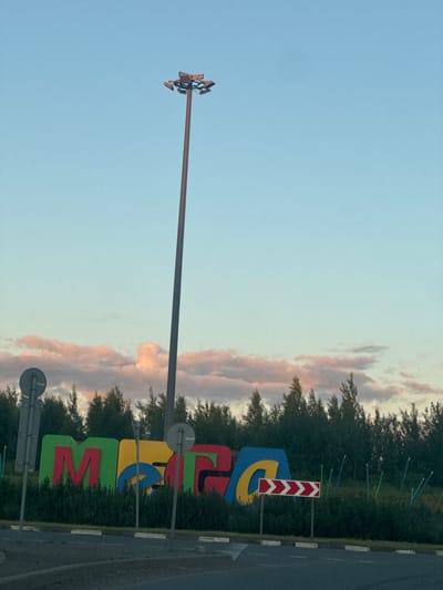 The image captures the MEGA shopping center's outdoor signage in Korabsel'ki, Russia, at dusk. The sign features large, colorful, three-dimensional letters spelling out "MEGA." The letters are a mix of green, red, yellow, and blue, with red "M," yellow "E," and blue "G" and "A" characters. The letters are set against a backdrop of dense green bushes. To the left of the sign, a tall lamppost with multiple lights is visible, its pole a reddish-brown color. Two circular road signs, one gray and one with a red border, are also present to the left and right of the main sign, respectively. A red and white chevron directional sign is positioned to the right of the "MEGA" sign. The sky above is a soft gradient of light blue and pale pink, indicative of sunset, with scattered, wispy clouds tinted pink. A line of dark green coniferous trees forms the horizon behind the signage and bushes. The foreground shows a portion of a paved road and a curb. No people are visible in the frame.
