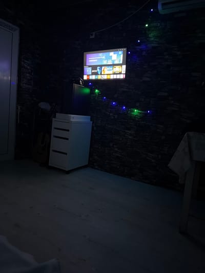 The scene depicts a dimly lit interior room in the city of Kermen, Bulgaria. The primary light sources are a wall-mounted smart television displaying a brightly lit interface with various application icons, and a string of blue and green LED lights strung horizontally across the stone-patterned feature wall. An air conditioning unit is mounted on the wall above the television. Below the television, a black rectangular object, possibly a speaker or subwoofer, rests on a white, four-drawer rolling storage unit. A light-colored acoustic guitar leans against the wall behind the storage unit. To the far left, a white door and door frame are partially visible. To the right, a table draped with a white, patterned tablecloth is present. The floor is light-colored wood laminate. No individuals are present, and no actions or interactions are depicted.