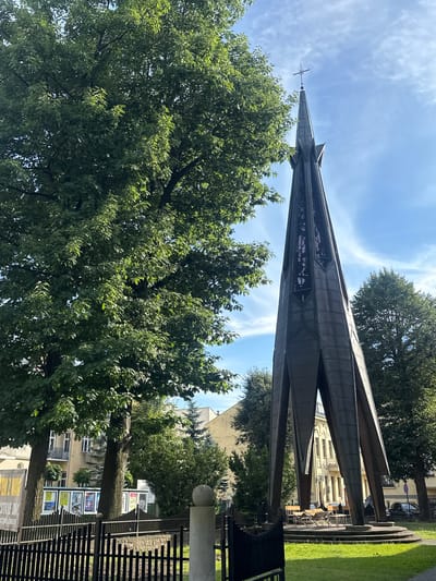 In Nowy Sącz, Poland, a tall, dark, metal spire with a cross at its apex stands prominently in an urban park. The spire's design is angular, with four large, angled legs supporting the tapering structure. Beneath the spire, a circular platform is visible with several chairs and tables, suggesting an outdoor seating area. Lush green trees, some with broad canopies and others with more slender forms, surround the spire and the park. In the background, traditional European-style buildings with light-colored facades and multiple windows are visible, interspersed with more trees. A black metal fence runs along the front of the park, and a sign with text in Polish is partially visible on the left side of the frame. The sky above is a clear blue with scattered white clouds. The scene appears to be set on a bright, sunny day.