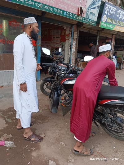 This image captures an outdoor street scene in Kaliganj, Bangladesh, specifically near the "Mosque Market" and a "ROYAL EXPRESS" business, as indicated by Bengali and English signage displayed above various shopfronts.
In the foreground, two adult males are prominent. On the left, a bearded man wearing a white patterned cap, a long-sleeved white kurta with decorative embroidery, white pants, and dark brown open-toed sandals stands, facing towards the right. On the right, a second adult male, also bearded and wearing a white skullcap, is dressed in a dark red long robe with white visible at the cuffs or hem, and light brown open-toed sandals. He is positioned with his back mostly towards the viewer, leaning over the seat of a black motorcycle with red accents on the fuel tank. A second black motorcycle is parked immediately behind the first.
The ground surface is unpaved, consisting of light brown earth or dirt with scattered debris, including fragments of pink and white plastic and dried organic matter. The background features several closed or partially open shopfronts, consistent with a commercial street or market area. The scene is illuminated by natural daylight. A timestamp of "04/12/2025 14:07" is present in the lower right corner of the image.
