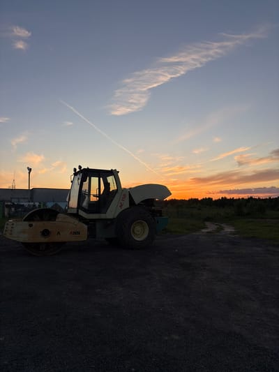 In the city of Baranovo, Russia, a roller compactor, identified by the markings "VOGELE" and "AC 150," is parked on a dirt path during sunset. The sky displays a gradient of colors, from pale blue at the top to vibrant orange and yellow near the horizon, with scattered wispy clouds and a contrail from an aircraft streaking across the upper portion of the frame. In the background, a line of trees forms a dark silhouette against the colorful sky. To the left of the compactor, industrial structures and a tall pole with lights are visible. The ground is covered in dark gravel. No people are present in the image.