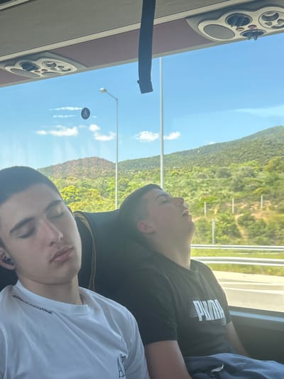 Two people are seated on a bus. One wears a black Puma shirt and the other a white Armani Exchange shirt. Both are asleep. The bus travels along a road, with a view of forested mountains visible outside the window. A street lamp and a sign with the Greek letter omega are visible through the window as well. The vehicle appears to be traveling through Vrasna, Greece. The interior of the bus is visible with AC and speaker.