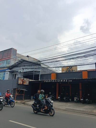 A street scene shows a gray, cloudy sky above buildings and numerous power lines. A "Profesional Gym" is visible, its entrance fronted by parked motorcycles. In the foreground, motorbikes are actively being driven on the street. One motorbike carries three people, including a child. The overall atmosphere suggests a bustling, ordinary day in an urban area.