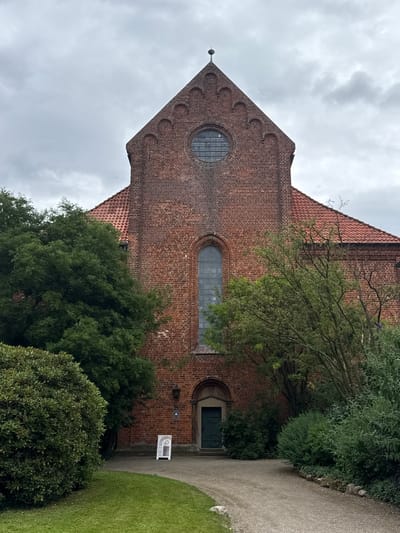 The image shows the exterior of the St. Mary's Church in Bassum, Germany. The church is constructed of red brick and features a steep, tiled roof. A large, arched window is visible on the facade, as well as a circular window above it. A smaller, dark green door is located at the base of the building. The church is surrounded by lush green trees and bushes. A gravel path leads to the entrance, and a small sign stands near the path. The sky is overcast with clouds.