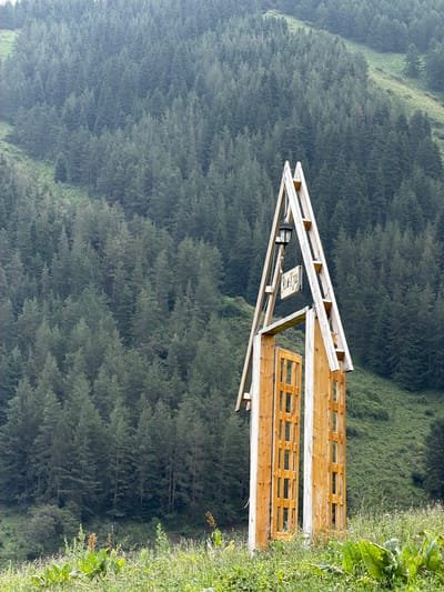 The image depicts a wooden structure, possibly a small shelter or viewing platform, set against a backdrop of a dense, evergreen forest covering a steep hillside. The forest extends into the distance, shrouded in a light mist or haze, creating a sense of depth and atmosphere. The wooden structure itself is constructed from light-colored planks, with a sloped roof and a latticed railing. A black lantern hangs from one of the roof supports, and a small, weathered wooden sign is suspended by chains from the structure. The sign has text on it, but it is not entirely legible due to the image quality and angle. The foreground on the right side of the image shows lush green vegetation, suggesting an outdoor, natural setting. The overall mood is serene and slightly mysterious, perhaps indicating a remote or natural tourist spot. The time of day appears to be daytime, with soft, diffused light, and the weather seems calm, possibly humid given the mist. The location context provided as Kaindy, Kyrgyzstan, suggests a mountainous, forested region.