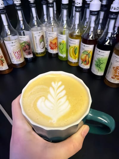A close-up, high-angle shot captures a light-skinned hand holding a white, faceted ceramic mug with a teal handle, filled with a frothy, light brown beverage topped with intricate white latte art depicting a stylized leaf. The mug is held in the lower central part of the frame. In the background, arranged on a dark, reflective surface, is a semicircular display of approximately eighteen clear glass bottles. Each bottle is fitted with a white pump dispenser and contains various colored syrups. The labels on these bottles display the brand "PINCH & DROP" and feature illustrations of different flavors such as popcorn, almonds, coconut, maple, cactus, banana, pear, mint, and gingerbread, with accompanying text in Cyrillic script. The scene is set in what appears to be a cafe or beverage preparation area in Lubnya, Russia.