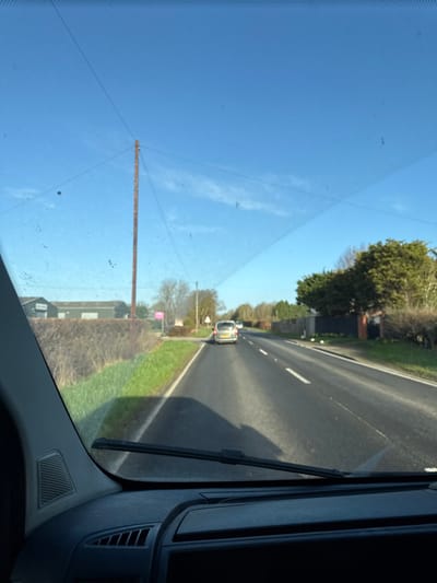 The image depicts a forensic view from the interior of a vehicle, facing forward on an asphalt road under a clear, bright blue sky with sparse, wispy clouds. This scene is located on an unspecified rural road within the Borough of Rushcliffe, United Kingdom.
In the immediate foreground, the top of a vehicle's dashboard is visible, featuring a speaker grille on the left and a portion of the windshield wiper blade at the bottom center. The windshield is visible, displaying accumulated dirt/dust and a faint pattern of cracks or scratches.
The road ahead is a single-carriageway with two lanes, marked by white dashed lines. A silver or light-colored minivan or MPV is observed in the distance, traveling away from the viewer, with another less distinct vehicle further behind it. Shadows cast by the vehicles are visible on the road surface, indicating sunny conditions.
To the left of the road, there is a grassy verge leading to a dense, trimmed hedge. Behind the hedge stands a tall, dark-colored utility pole with multiple overhead wires extending across the sky. Further back and to the left, several large, dark grey industrial or agricultural buildings, possibly barns or sheds, are present. Near a small track or entrance on the left, a distinct pink sign, likely a real estate advertisement, is visible alongside a white triangular road sign with a red border and an 'X' symbol, indicating a crossroads or junction ahead.
To the right of the road, a similar grassy verge abuts a continuous line of dense trees and hedges, which partially obscure any structures beyond them. No people are discernible within the frame.