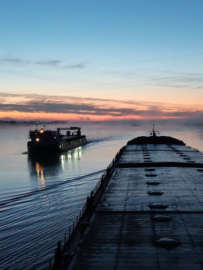 The image depicts two barges on a river at dawn or dusk. The foreground barge is large, flat, and gray, with a series of oval hatches visible on its surface. A second barge is seen further away, illuminated with several lights reflected on the water. A misty horizon is illuminated with an orange and pink glow, transitioning to a blue sky. Power lines are strung overhead. The photograph was taken in Gari, Russia.