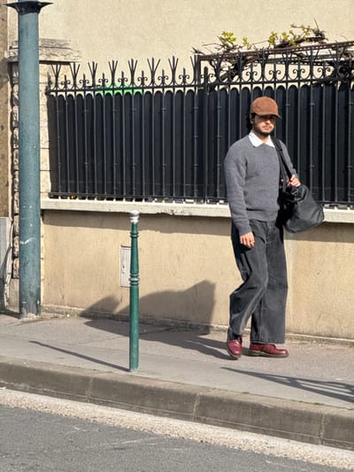 The image depicts a lone adult male walking on a concrete sidewalk in Asnières-sur-Seine, France. The individual, centrally positioned and facing right, is captured in motion. He is dressed in a brown corduroy baseball cap, dark sunglasses, a white collared shirt beneath a gray crew-neck sweater, dark gray wide-leg jeans, and dark red lace-up boots. A black duffel bag is carried over his left shoulder. To the left, a green ornamental bollard with a polished cap stands on the sidewalk, casting a long shadow across the pavement. A dark green utility pole is partially visible on the extreme left. The background features a continuous light beige stucco wall, surmounted by a black wrought iron fence with decorative spiked finials. Above the fence, minimal green foliage is present. A small, light-colored electrical junction box is affixed to the stucco wall behind the bollard. The immediate foreground includes a dark asphalt road bordered by a concrete curb. The scene is brightly lit by direct sunlight, producing sharp, elongated shadows on the ground.