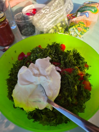 The image displays a high-angle view of food preparation taking place in Motyakovo, Russia. In the foreground, a bright lime-green plastic bowl contains a salad composed of finely chopped green herbs, including dill and cilantro, mixed with diced red tomatoes. A large dollop of white, creamy dressing is situated centrally in the salad, with a silver spoon partially submerged and positioned diagonally, indicating that the dressing is being stirred into the ingredients.
In the background, to the left, a reddish-brown jar with a gold lid is visible next to a clear, empty drinking glass. A person's hand and forearm are partially visible at the upper left. Towards the center, a clear glass with a dark, wave-pattern design stands alongside a small white bowl containing several small, round red items. Further back, two plastic bags are present: one appears open and contains chopped green vegetables, while the other is a sealed package of bread, clearly labeled with Cyrillic text including "Батон" (Baton), and features green and yellow packaging elements. The entire scene is set on a light blue or green surface, illuminated by bright natural light.