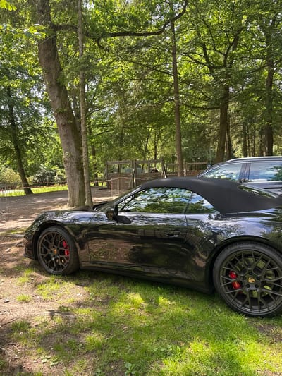 The image depicts a black Porsche 911 convertible, likely a 992 generation, parked on a grassy verge adjacent to a dirt path in a densely wooded recreational area. The car's black soft top is raised. It features dark multi-spoke wheels with visible red brake calipers, indicating high-performance brakes. Pirelli tires are mounted on the wheels. Sunlight filters through the overhead tree canopy, casting dappled light and shadows across the scene and reflecting off the car's glossy bodywork.
In the mid-ground, a dirt or gravel path is visible, bordered by a temporary barrier consisting of metal posts and red-and-white striped tape. Beyond this barrier, towards the left, are more trees and green foliage, including what appears to be rhododendron or similar flowering shrubs. Towards the right background, behind the Porsche, a dark-colored SUV or wagon is partially visible, parked. Further back in the center-right, large, rustic wooden structures, consistent with a playground or an adventure course, are present. A faint human figure is discernible sitting on a distant wooden bench or structure behind these wooden elements. The ground consists of patches of green grass interspersed with exposed soil and leaf litter.
The scene suggests a **wooded recreational area or a 'Waldspielplatz' (forest playground) within the city limits of Oberndorf am Neckar, Germany**.