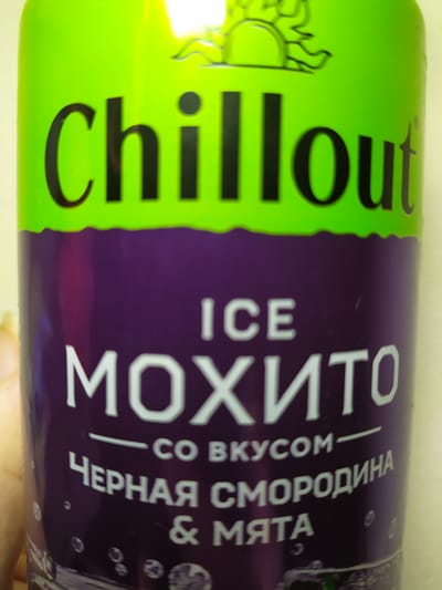 A close-up, vertically oriented shot focuses on a bright green and purple beverage can, likely a soda or energy drink. The can is tilted, with the lighting suggesting an indoor setting, possibly a brightly lit room or under studio lights. The background is a blurred, neutral color.

The green portion of the can features the word "ChillOut" in large, bold, black lettering. To the left of the text, there's a stylized graphic of a sun or flower, also in black. The purple section of the can is dominated by white text. At the top, "ICE" is written horizontally. Below that, the word "МОХИТО" (Mojito) is prominently displayed vertically in large, white, sans-serif letters. Underneath, smaller text reads "— СО ВКУСОМ —" (— WITH TASTE —). Further down, another line of text reads "ЧЕРНАЯ СМОРОДИНА" (Black Currant). The bottom line of text is "& МЯТА" (& Mint). Small, circular bubbles are visible on the purple background, suggesting the carbonation of the drink. The overall impression is one of a refreshing, flavored beverage. No people are visible in the frame.