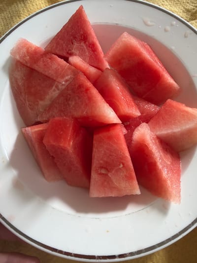 A white plate with a double-banded black and grey rim is filled with chunks of cut watermelon. The watermelon is a deep pinkish-red color, and some pieces have a visible, watery sheen. The plate is placed on a yellow textured surface. A finger is visible in the bottom right corner of the image. The image appears to be taken indoors under natural lighting. There is no indication that the image was taken in Pentre, United Kingdom.