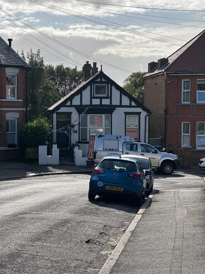 This image depicts a residential street scene in Thanet, United Kingdom. The central focus is a two-story mock-Tudor style house, number 48, featuring white stucco walls with dark timber framing and a prominent front gable. The house has a dark front door, ground-floor bay windows with visible red/orange interior curtains and criss-cross pane patterns, and a smaller window in the gable. White decorative low walls flank the front path, and a large white spiderweb Halloween decoration is affixed to the house near the entrance.
To the left of the central house is a red brick residence with white window frames and visible white shutters. To the right is another red brick house with white window frames. Overhead power lines are visible against a partly cloudy sky.
In the foreground, a blue Mazda hatchback (UK registration LV67 GLK) with a "UK" sticker on the rear is parked angled towards the curb on the asphalt road, which shows minor wear and potholes. Behind the blue Mazda, a silver/grey pickup truck with a white enclosed utility canopy and partially visible text "Management" on its side is parked. A third white vehicle is partially visible behind the pickup on the far right. No individuals are present in the scene.