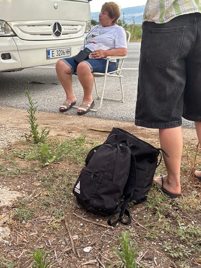 The image depicts a scene on the side of a road, likely in or near Prosotsani Municipal Unit, Greece, given the presence of a Bulgarian license plate on the vehicle. In the foreground, two black backpacks are placed on the ground next to scattered dry vegetation and some small green plants. One backpack, an Adidas brand, is in sharper focus. To the right of the backpacks, the lower legs and feet of a person wearing dark denim shorts and black flip-flops are visible. Further back, a woman sits on a white folding chair beside a white Mercedes-Benz bus. She is wearing a white t-shirt and denim shorts and is holding a clear plastic bottle. Another person, partially visible from the waist down and wearing a plaid shirt and dark denim shorts, stands to the right, their back to the camera. The bus has its headlights on, and a license plate reading "BG E 3206 HH" is clearly visible. The ground is a mix of asphalt and dirt, with sparse vegetation. The sky in the background is a light blue with some wispy clouds.