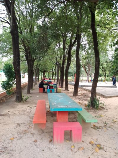 The image depicts an outdoor park setting, likely in Villa Hayes, Paraguay, characterized by tall trees providing shade over colorful concrete picnic tables and benches. The ground is sandy with scattered leaves and debris. One person is sitting at a table further down the path, and two people are standing and talking in the background near a playground. The foreground table is a vibrant combination of turquoise, red, and green. The scene suggests a public recreational area.