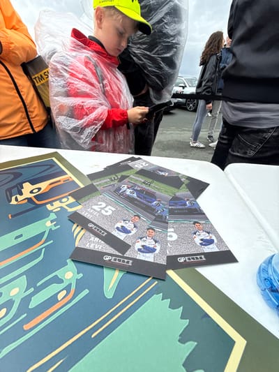 This image captures an outdoor scene at the Sachsenring circuit in Hohenstein-Ernstthal, Germany, under an overcast sky, indicating inclement weather. In the foreground, a white table displays promotional materials for a motorsport event. On the left, a large poster features stylized racing graphics in blue, green, and orange. To the right, a stack of smaller flyers showcases race car driver Ariel Levi, number 25, in a white racing suit, alongside a blue race car on a track, all branded with "GP ELITE." A partial blue, ribbed container is visible on the far right of the table.
In the midground, a young Caucasian boy, approximately 8-12 years old, is centered. He wears a bright yellow baseball cap with "B. MANTHEY" text and a clear plastic rain poncho over a red jacket. He is intently focused on a small object, possibly one of the flyers, held in his hands. To his immediate left, the partial torso of an adult in an orange jacket with a black shoulder bag displaying "TM SMART" text is visible, also wearing a rain poncho. Further back and to the boy's right, other individuals are partially visible, including one person wearing light grey jeans and white Adidas sneakers with black stripes. In the background, on an asphalt surface, a white car is partially visible, consistent with a motorsport paddock or fan zone environment. The presence of multiple individuals in rain ponchos confirms rainy conditions.