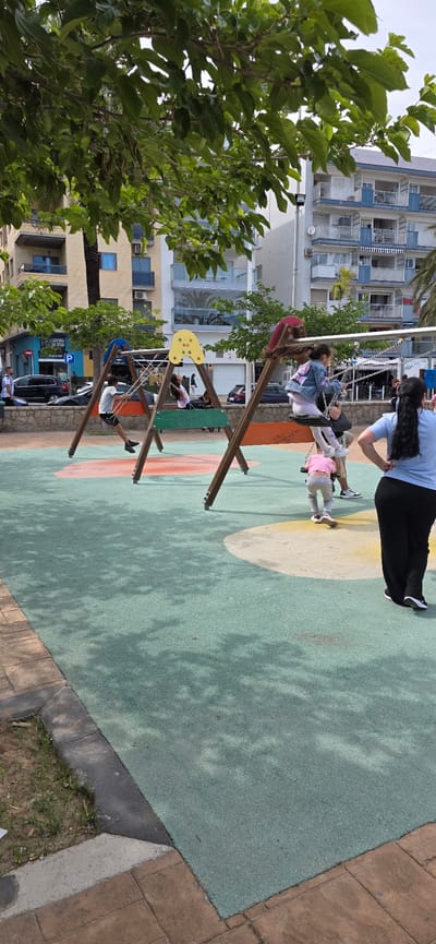 A horizontal outdoor shot depicts a playground in El Port De Sagunt, Spain.
A swing set is present, occupied by two children and pushed by an adult. The playground's floor is a green and beige patterned surface with additional colored shapes near the swings. Adjacent to the playground is a tiled walkway.
Behind the playground, buildings with architectural variations stand. Trees, especially a large one on the left, frame the scene. Cars and other pedestrians are visible along the street behind the playground. The lighting indicates daytime.