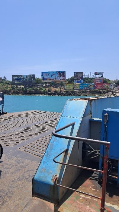 The image presents a daytime scene from the deck of a ferry traversing the Likoni Channel in Mombasa, Kenya, looking towards the Ganjoni/Shimanzi ward. In the foreground, a heavily worn, dark-colored metal deck surface with a repeating raised pattern is visible. To the right, a rusted, blue-painted metal structure with an angular, sloped top section stands. Attached to this structure is a three-tiered, weathered metal handrail, exhibiting rust and red paint residue. A portion of a bicycle wheel, including a black tire and silver rim, is visible on the far left edge of the deck.
The midground features a wide expanse of clear, calm turquoise-blue water. The background reveals a vegetated coastline or landmass, rising gently from the water's edge. Positioned along the crest of this landmass are at least eight large billboards of varying sizes and designs. Advertisements visible include "quickie" on the far left billboard, and another large billboard featuring product bottles and individuals with the text "TUCANO". Other billboards display diverse colors and imagery, some appearing to be advertisement structures without specific content discernible. The sky above is clear and bright blue, devoid of clouds. No individuals or active interactions are visible.