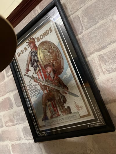 A framed vintage poster displays a patriotic scene. "U.S.A Bonds" is prominent, next to an image of the Statue of Liberty. Another figure, possibly a soldier or scout, wields a sword and shield. Text on the poster reads "Third Liberty Loan Campaign Boy Scouts of America" and "Weapons for Liberty." The poster's design includes elaborate embellishments, captured within a dark, ornate frame. The artwork is mounted on a brick wall.