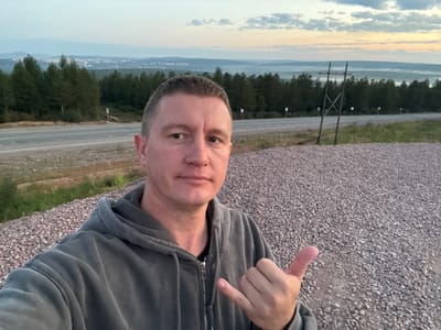 A man in a gray hooded sweatshirt is taking a selfie outdoors. He is standing on a gravelly surface with a highway and a dense forest in the background. The sky is overcast with hints of sunset colors. In the distance, a city can be seen. The man is making a "shaka" hand gesture. This location is identified as being near Serebryany Bor, Russia.