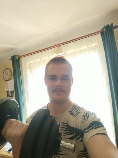 A young man with short brown hair and a mustache holds a black dumbbell in his right hand, performing a bicep curl. He is wearing a light-colored t-shirt with a dark pattern of leaves. The background features a window with sheer white curtains and dark green drapes, suggesting an indoor setting in a residential area. A round wall clock with black numerals and hands is visible on a yellow wall to the left. The lighting indicates it is daytime. The scene appears to be captured in a room within a home in Cēsis, Latvia, based on the context provided.