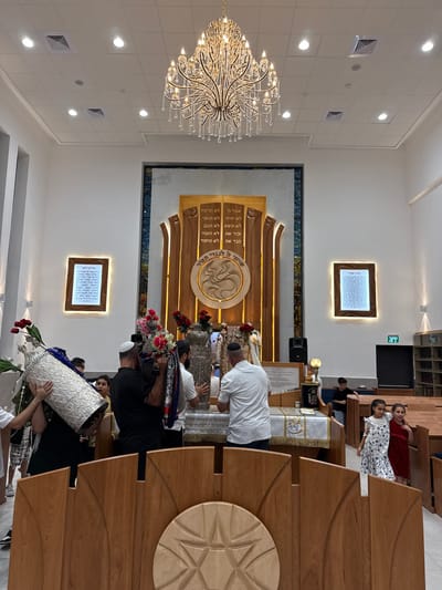 This image depicts the interior of a modern synagogue in Beer Yaaqov, Israel, during a religious ceremony involving Torah scrolls. The space features a high white ceiling with numerous recessed lights and a large, multi-tiered crystal chandelier. The far wall is dominated by an ornate wooden Torah Ark (Aron Kodesh) with carved panels, illuminated elements, and Hebrew inscriptions. The Ten Commandments are inscribed above a central carved motif, with "שויתי ה' לנגדי תמיד" (I have set the Lord always before me) inscribed below it. Two illuminated framed plaques with Hebrew text flank the Ark.

In the mid-ground, a group of approximately five adult males, most wearing kippahs, are actively engaged with Torah scrolls. One man on the far left carries a large, elaborately decorated silver-cased Torah scroll adorned with red roses over his shoulder. Another man, in a black shirt and white kippah, carries a similar silver-cased Torah scroll draped with a colorful *tallit* and decorated with pink and red flowers. A third man, in a white polo shirt and white kippah, is positioning a Torah scroll onto a low wooden table covered with a white and gold embroidered cloth, situated directly in front of the Ark. Another man is partially visible behind the table, also handling a scroll.

The foreground features a wooden partition with a carved Star of David motif. To the right, rows of wooden pews are visible, and two young girls in dresses are standing, smiling towards the viewer. A young boy in a black shirt is observed further back among the pews. On the far right wall, an illuminated green "EXIT" sign (יציאה) and bookshelves are present.