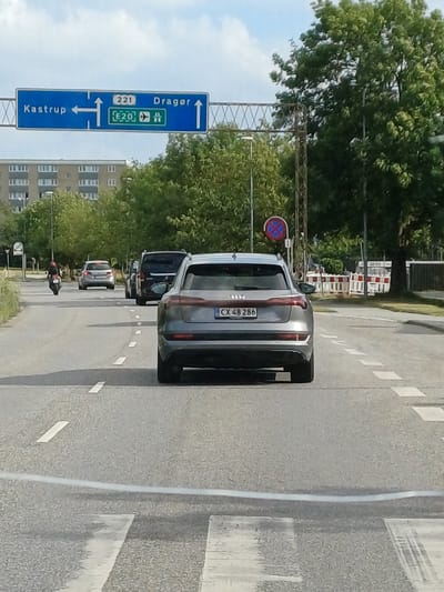 The image shows a street scene in Kastrup, Denmark, with several cars visible. The main subject is a silver car with license plate CX 48 286, seen from the rear. The car is on a road with white lane markings. To the left of the road, there is a blue directional sign with white text indicating "Kastrup" with arrows pointing straight and right, and "Dragør" with an arrow pointing right. The sign also shows "221" and "E20" along with an airplane symbol, suggesting proximity to the airport. A traffic signal with a red and blue circular sign showing a red diagonal line across a red circle is visible. In the background, trees and buildings line the street. Construction barriers are also present in the distance. The lighting suggests it is daytime, and the sky is partly cloudy. The scene appears to be a typical urban or suburban street.