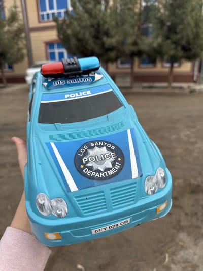 The image depicts a light blue plastic toy police car held by a person's hand in Apshak, Uzbekistan. The toy car features a red and blue siren light bar on the roof and decals stating "LOS SANTOS POLICE DEPARTMENT" on the hood, and "POLICE" and "LOS SANTOS" above the windshield. A license plate on the front bumper reads "01 Y 028 CB". A hand, possibly a child's, wearing a light-colored long-sleeved garment, supports the toy from the left. The background, out of focus, shows a dirt ground, several evergreen trees, and a multi-story building with a light brown facade and blue window frames. A white vehicle is partially visible behind the trees in the distance.