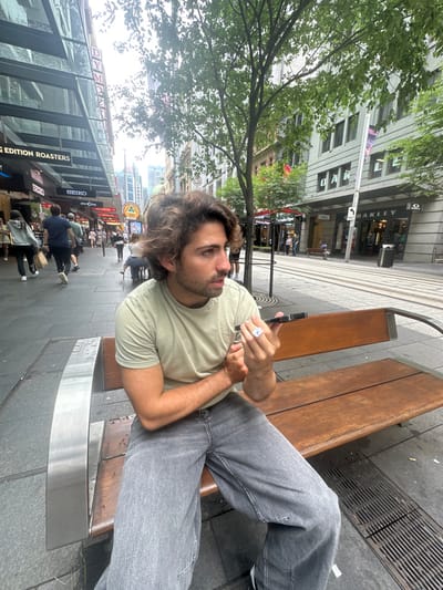 A man sits on a wooden bench outdoors, holding a small white object. He wears a light green t-shirt and gray jeans. The background reveals a city street lined with buildings, some obscured by green trees. A street sign and storefronts are visible, along with pedestrians walking along the sidewalk. The image appears slightly rotated.