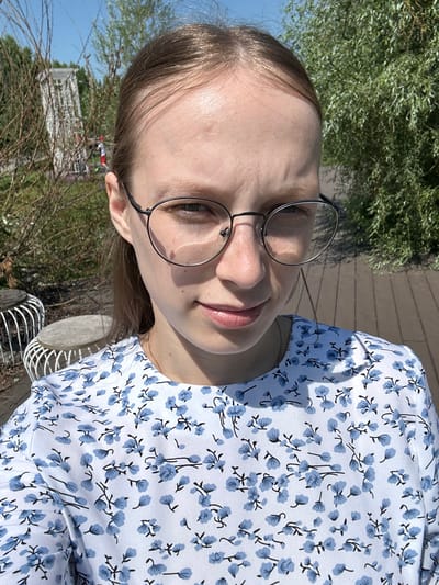 A young woman with fair skin, brown hair, and glasses is taking a selfie outdoors. She is wearing a white dress with a blue floral pattern. The background suggests a park or outdoor seating area with wooden planks on the ground, some bushes, and trees. In the distance, a white structure, possibly a gazebo or trellis, is visible. The lighting indicates it is daytime, with a clear blue sky.