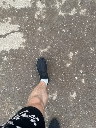 The image shows a first-person perspective of someone walking on a wet asphalt surface. The asphalt has patches of lighter colored material, possibly concrete or lighter asphalt, and is speckled with small stones and debris. The person is wearing a black athletic shoe with white laces and a white sock. A portion of their bare leg is visible above the sock, and they are wearing black shorts with a pattern of light pink cherry blossoms. The scene appears to be outdoors on a street or pathway, indicated by the urban ground texture. There is no information in the image to suggest it is in Oral, Kazakhstan.