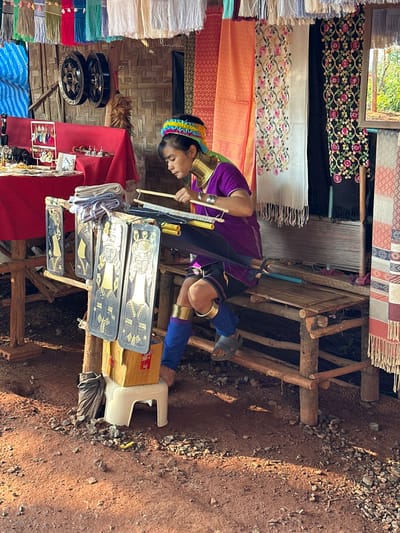 A Long Neck Karen woman weaves using a traditional loom outdoors. She wears coils around her neck and ankles, along with a colorful headpiece. Finished woven textiles hang behind her. A display of merchandise, including decorative plates, sits on a table in front of her. The setting appears to be a roadside shop or market stall with gravel and red soil visible.