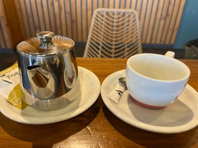 A silver teapot sits on a white saucer to the left of a white coffee cup on a white saucer. The coffee cup is empty. A silver teaspoon lies on the saucer, next to a white sugar packet. The scene is likely inside a cafe or restaurant, as a wooden table and an empty chair can be seen in the background. The scene is located at the coordinates 43.321, -2.009 in the city of Donostia-San Sebastián, Spain.
