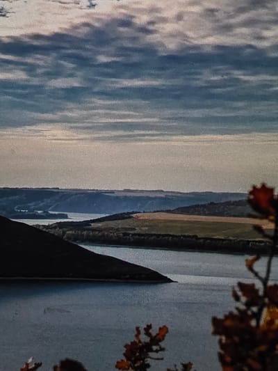 This is a landscape photograph featuring a wide body of water, likely a river or reservoir, winding through rolling hills. The foreground is dominated by a dark, steep hillside on the left, casting a shadow over the water. The middle ground displays a textured landscape with patches of greenery and cultivated fields, dissected by the watercourse. In the distance, layers of hills fade into a hazy horizon. The sky is filled with a dramatic, cloudy pattern of blues and grays, with streaks of lighter clouds suggesting diffused sunlight. The overall impression is one of a natural, serene environment, possibly captured during autumn given the hints of warm colors in the foliage on the right. The location is identified as being in the vicinity of Dubno, Ukraine. There are no people or animals visible in the image.