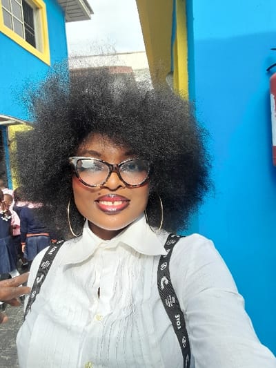 A Black female adult is depicted in a close-up, chest-up portrait shot, positioned outdoors in Ajah, Nigeria. She has dark, voluminous afro-style hair, is wearing large tortoiseshell-patterned eyeglasses with white accents, and gold hoop earrings. Her lips are adorned with dark, glossy lipstick, and she is smiling, exposing teeth. She wears a white collared shirt with vertical pintucks on the front and black suspenders featuring white text/logos, including "TOAE." In the background to her left, several children in school uniforms (white shirts and blue skirts/trousers) are partially visible and out of focus. The immediate background consists of brightly colored building walls: a dominant blue wall on the right with a red fire extinguisher mounted on it, and another blue wall on the left featuring a yellow-framed window. A yellow architectural trim element is also visible. A hand, possibly belonging to another individual, is partially visible in the lower-left foreground.