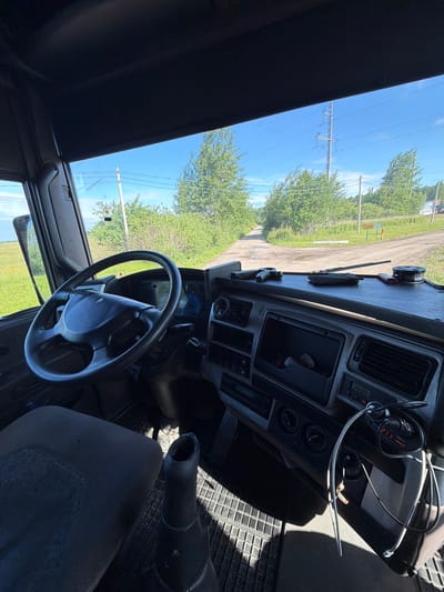 This media file shows the interior of a truck cabin, looking out at a rural, daytime scene. The truck appears to be parked on a dirt road, with lush green trees and vegetation lining the sides. In the background, power lines and poles are visible against a clear blue sky, suggesting good weather. The interior of the truck is functional, with a steering wheel, dashboard, and car accessories like a phone charger and a digital display showing "27.1". There are no people visible in the image. The overall impression is one of a vehicle stopped in a peaceful, outdoor environment, likely during the summer months due to the foliage. The location context of Zarech'e, Russia, adds to the rural setting.
