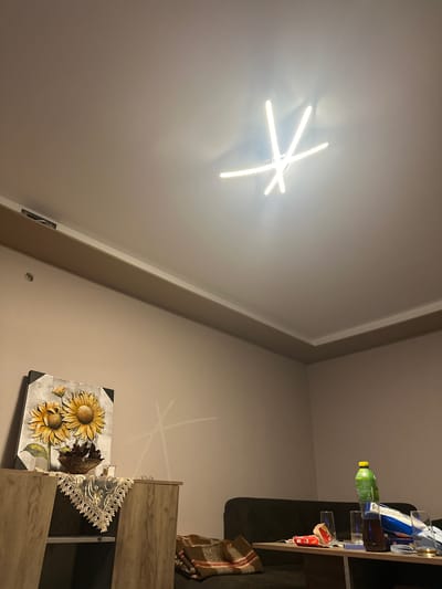 The image depicts the interior of a residential room in Krupnik, Bulgaria, likely a living area. The ceiling is white, featuring a modern, multi-bar LED light fixture illuminated in a star-like pattern. Beige-colored walls extend below a white and brown cornice, with an exposed or unfinished section visible in the cornice on the left wall.

On the left, a light wood-grain cabinet or credenza supports a framed canvas artwork depicting two large yellow sunflowers. A beige, embroidered lace doily is draped over the cabinet's corner. Other items on the cabinet include a bronze-colored bowl containing dark foliage, a small glass candle holder, and a pair of eyeglasses.

In the center-right, a dark-colored sofa is partially visible. In front of the sofa is a dark coffee table laden with several items: a green plastic bottle with a yellow cap, a brown glass bottle, three clear drinking glasses (two appearing partially used), a white and blue rectangular package, a red package with white lettering, and a white brush. A plaid blanket in brown and orange tones is draped over the left side of the sofa. No individuals are present in the scene.
