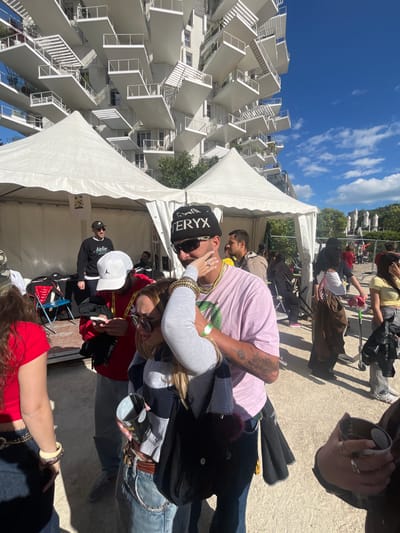 The image captures an outdoor scene in Montpellier, France. The background features a distinctive modern building with numerous white balconies jutting out, partially obscured by a large white tent. Several individuals are visible; a man in a pink shirt and a black beanie with white text is embracing a blonde woman in a grey long sleeve shirt with jewelry on both wrists. Near them, a man with a white baseball hat and black sweatshirt stands, while other people are seated or standing nearby. They are on a paved area with more people in the background.