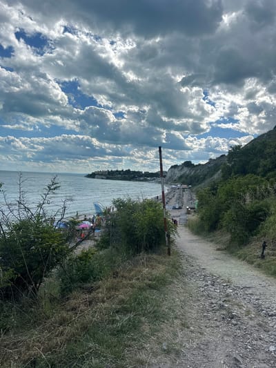 The image depicts a scenic view of a beach in Topola, Bulgaria, under a dramatic sky filled with clouds. In the foreground, a gravel path leads down towards the beach, bordered by dry grass and dense green foliage. A red and white striped pole stands near the start of the path.
The beach itself is populated with numerous beach umbrellas and lounge chairs, suggesting a popular tourist spot. People are visible on the sand, some relaxing under umbrellas, others near the water. The sea is calm with gentle waves, and a long stretch of coastline with buildings and a white cliff face can be seen in the distance. The overall atmosphere is one of a bright, slightly overcast day at a seaside resort.