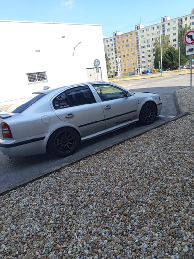 The image shows a silver Skoda Octavia car parked on a gravel surface next to a paved area. The car has tinted windows and custom alloy wheels. There are stickers on the rear window, one of which reads "GIRL RACER". The car appears to be modified with a lowered suspension.
In the background, there is a tall residential building with multiple floors and many windows. The building is light-colored, with some sections appearing to be a yellowish-brown hue. A street lamp is visible in front of the building. Further in the background, there are trees and more buildings. A traffic sign with a red circle and a black arrow indicating a left turn is visible at the top of the image, along with a sign depicting a truck.
The scene suggests daytime with clear skies, as indicated by the bright lighting and the blue sky. The car is parked at an angle, making it the primary subject of the image. The setting appears to be a residential area or parking lot in Zvolen, Slovakia, given the context provided. There are no people visible in the image. The overall mood is neutral, focusing on the car and its surroundings. The gravel area and the building suggest an urban or suburban environment.
