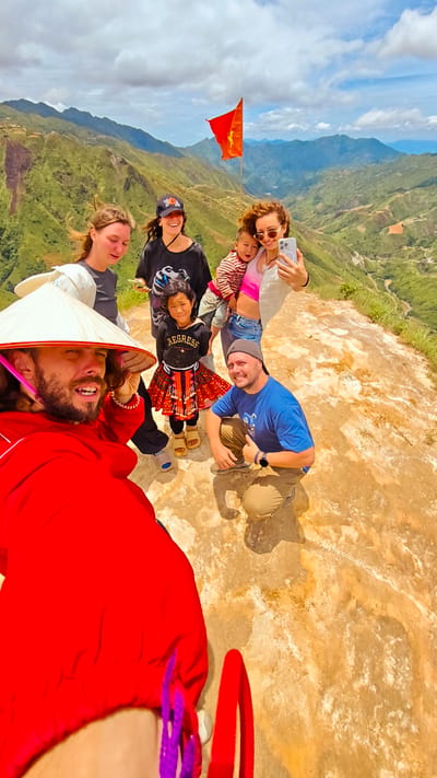 In the city of Bắc Yên, Vietnam, a group of six people are gathered on a rocky, elevated viewpoint overlooking a vast, green mountain landscape with terraced fields. The sky is cloudy with patches of blue. A Vietnamese flag is prominently displayed on a pole.

In the foreground, on the left, a man with a beard wears a red hooded jacket and a conical hat, with his face partially visible as he holds a phone, likely taking a selfie. To his right, a woman with curly hair, wearing a white tank top and sunglasses, holds a baby. She is taking a selfie with her phone. Behind her, a young girl in traditional attire, with a black hoodie that reads "REGRESS" and a colorful patterned skirt, stands looking at the camera.

To the left of the young girl, a woman in a black t-shirt and a black baseball cap smiles. Next to her, a woman in a grey t-shirt and white jacket is partially visible. On the right side of the frame, a man in a blue t-shirt and a dark baseball cap is kneeling and giving a thumbs-up gesture. The ground they are standing on is a rough, light-colored rocky surface with some brown patches. The overall scene captures a moment of travel and enjoyment of the scenic vista.