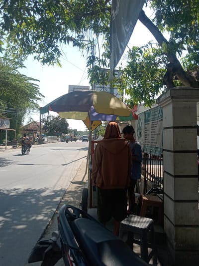 A sunny daytime scene on a street in Sukowati, Indonesia. A large, multi-colored umbrella shades two individuals standing near a fence. One person, wearing a brown hooded sweatshirt and dark pants, stands with their back to the camera, facing towards the street. The second person, a young male in a blue shirt and dark pants, stands to the right of the first person, looking at them. Behind the fence, a white banner with green lettering is attached to a wall. To the left of the umbrella, a motorcycle is parked on the sidewalk, with its rear visible. The street is asphalted and appears to be a public road with ongoing traffic. In the distance, more vehicles and buildings are visible, along with lush green trees lining the roadside. The overall atmosphere suggests a commercial or residential area with pedestrian and vehicle activity.