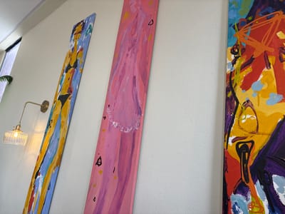 Three abstract paintings hang vertically on a white wall. The painting on the left is a riot of bold colors, featuring swirling brushstrokes of purple, yellow, red, and blue. The middle painting is predominantly pink with lighter pink and purple accents. Some black marks and yellow dots are scattered across its surface. The painting on the right is a mix of blues, yellows, reds, and black. A brass wall sconce with a clear, ribbed glass shade holding a lit bulb is mounted on the wall between the middle and right paintings, casting a warm glow. The paintings appear to be hung in a modern interior space, possibly an art gallery or a well-decorated home. The light from the sconce and possibly natural light from a nearby window suggest it could be daytime or early evening. There is no visible text in the image.