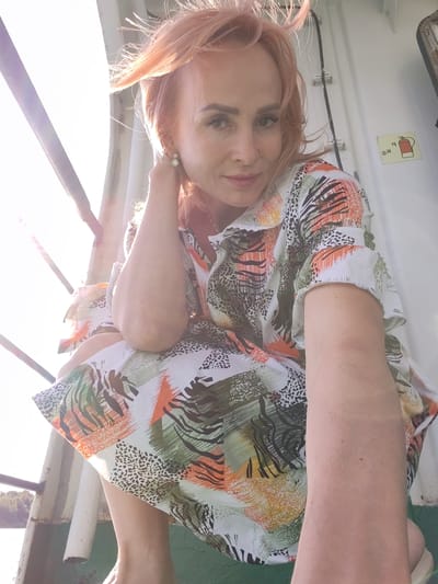 This media file shows a woman with light pink hair, wearing a colorful dress with an abstract print. She is looking at the camera with a slight smile, her left hand touching her neck. The background appears to be outdoors, possibly on a boat or ferry, with a railing and a white structure behind her. Sunlight is coming from the left, creating a lens flare and illuminating her hair and face. A sign with a fire extinguisher symbol is visible on the white structure. The scene suggests a casual, daytime setting with warm weather. The location context provided is Nova, Russia.