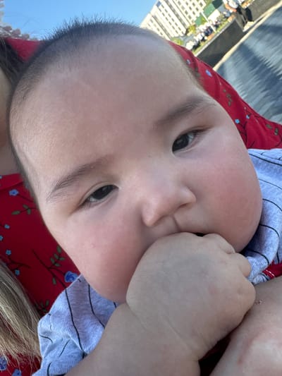 A close-up, head-and-shoulders shot captures a baby's face, with the majority of the frame occupied by the child's head and a portion of their body. The baby's right hand is brought up to their mouth, with fingers partially inserted. The baby is wearing a gray, striped shirt. Behind the baby, a red fabric with a floral print is visible, along with a strand of blonde hair. The background is slightly out of focus, showing a blue sky and buildings that appear to be in an urban setting, possibly Manas, Kyrgyzstan. The lighting suggests it is daytime, with the sun casting shadows.